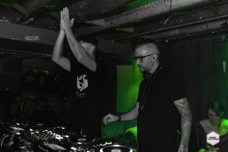 A DJ claps above the decks as a friend looks on. Launch night, May 2025