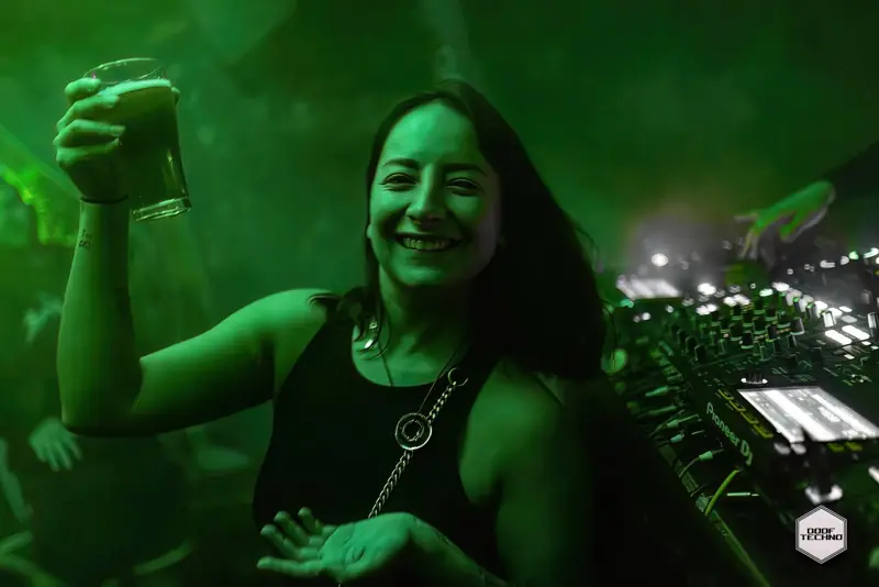 A dancer raises a glass and grins beside the booth. Launch night, May 2025