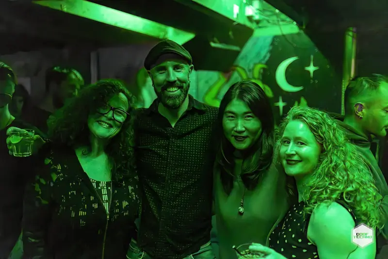 Four friends arm in arm, all smiles, under the moon mural. Launch night, May 2025