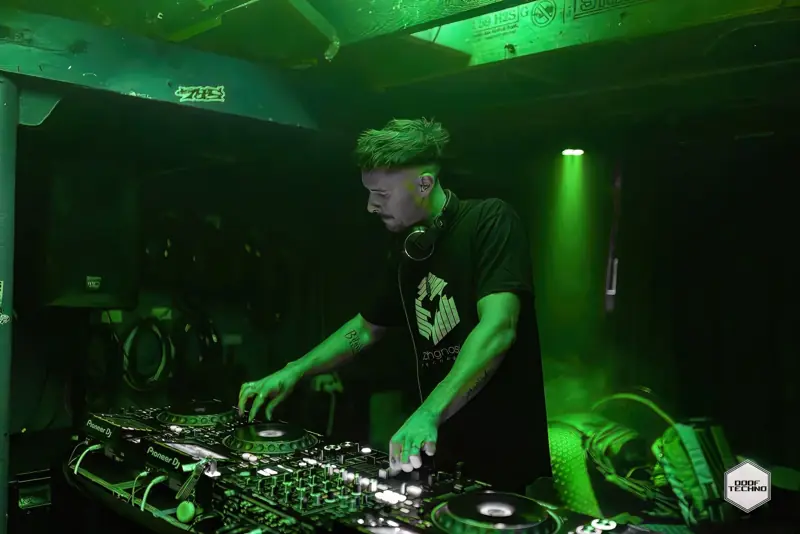 A DJ leans into the mixer, Techgnosis logo on his shirt. Launch night, May 2025